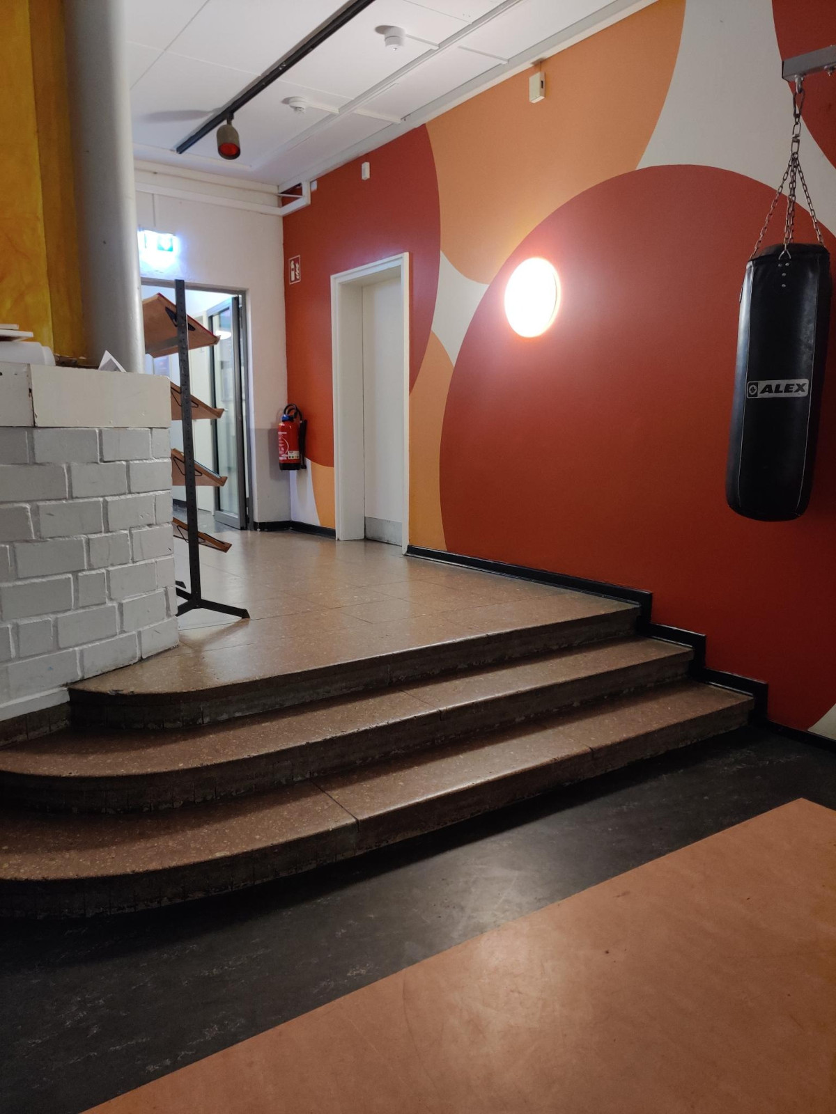 Blick aus dem Café-Bereich in Richtung Flur. Der Boden des Cafés ist niedriger als der Flur, und der Übergang besteht aus drei Stufen (in üblicher Treppenstufen-Höhe), die über die ganze Breite des Flurs gerade sind. An der linken Seite, wo sie überstehen und ins Café hineinragen, sind sie abgerundet. An der Wand neben den Stufen gibt es kein Geländer.