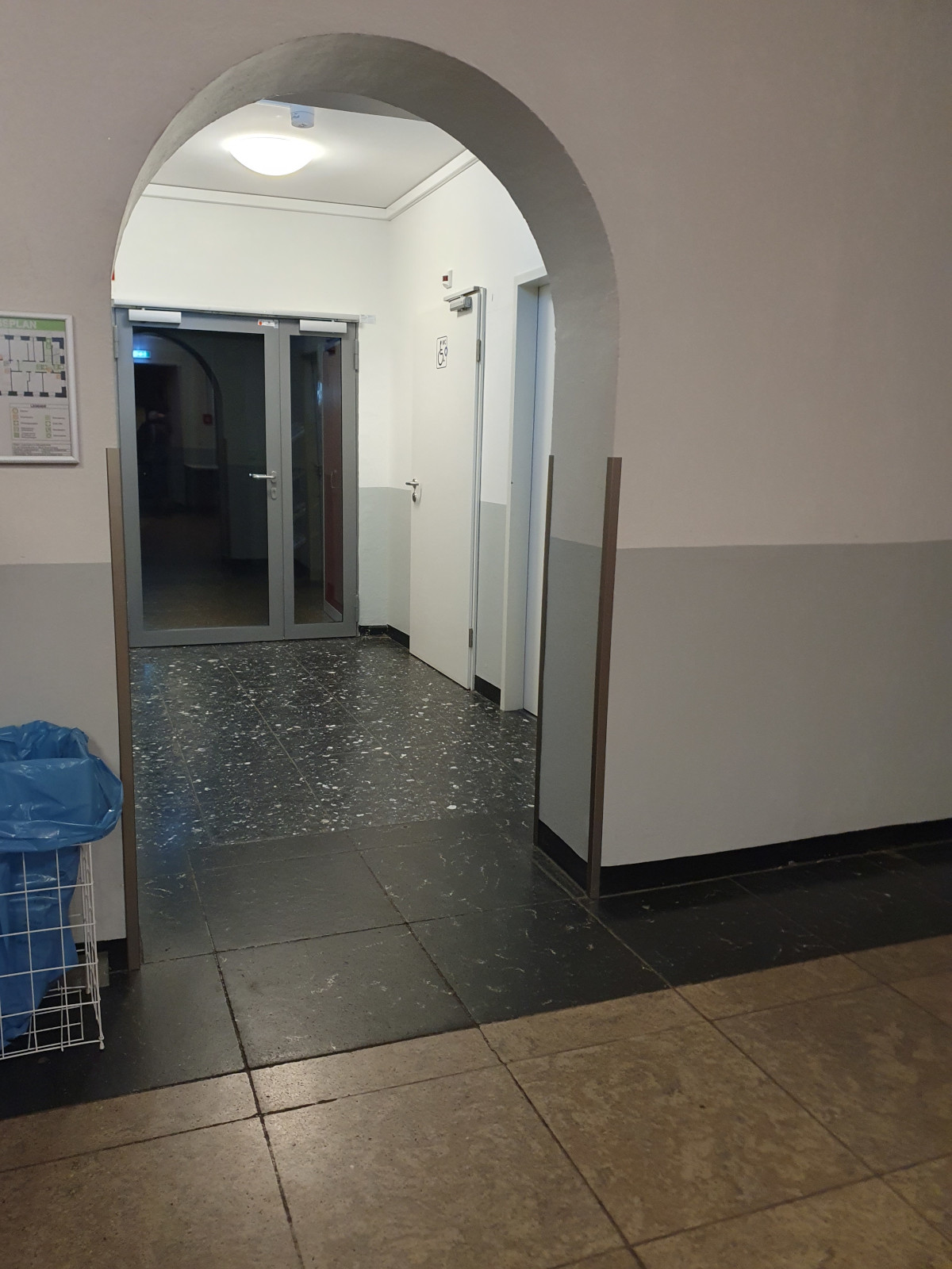 Blick von der Eingangstür durch den Flur im Erdgeschoss. Auf der rechten Seite ist unter anderem eine Toilettentür mit Rollstuhl-Symbol zu sehen. Geradeaus ist die Glastür zum Café. Hinter der Glastür ist auf dem Foto allerdings nichts zu sehen, weil das Licht aus ist.